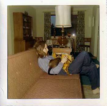 A young woman slouched on the couch, trying to knit with a Siamese cat draped across her lap.