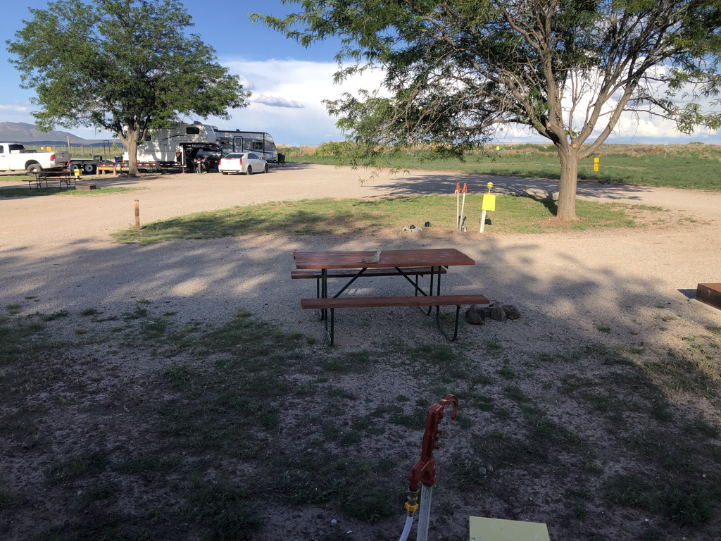 View of a campground with packed dirt sites. There is a picnic table in front, and some large shade trees beyond.