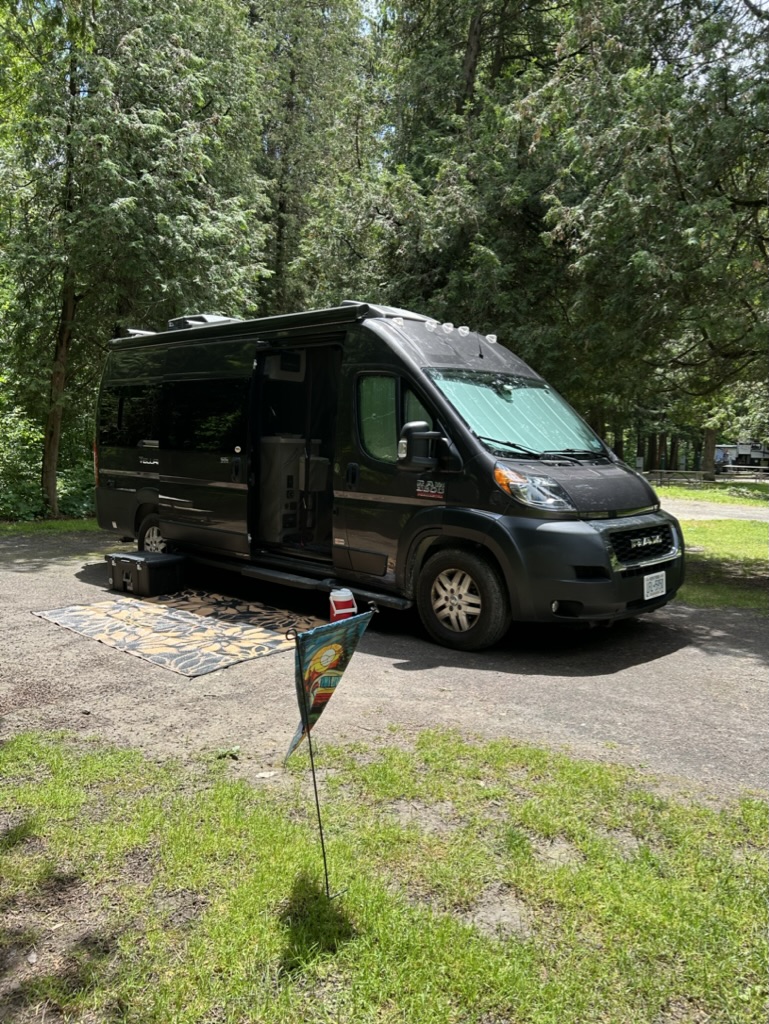 A charcoal gray camper van parked on a gravel pad with tall trees behind it and a camp flag waving in the breeze in front