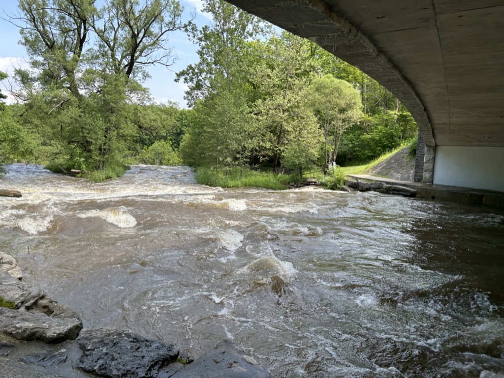 Brown, white-capped water flows under an overpass toward the top of a waterfall