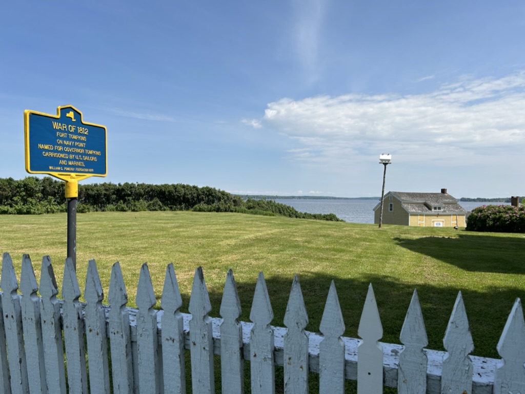 A green field with a white picket fence in front and a square building in the distance. The field overlooks a harbor, and a sign near the fence identifies it as Fort Tompkins