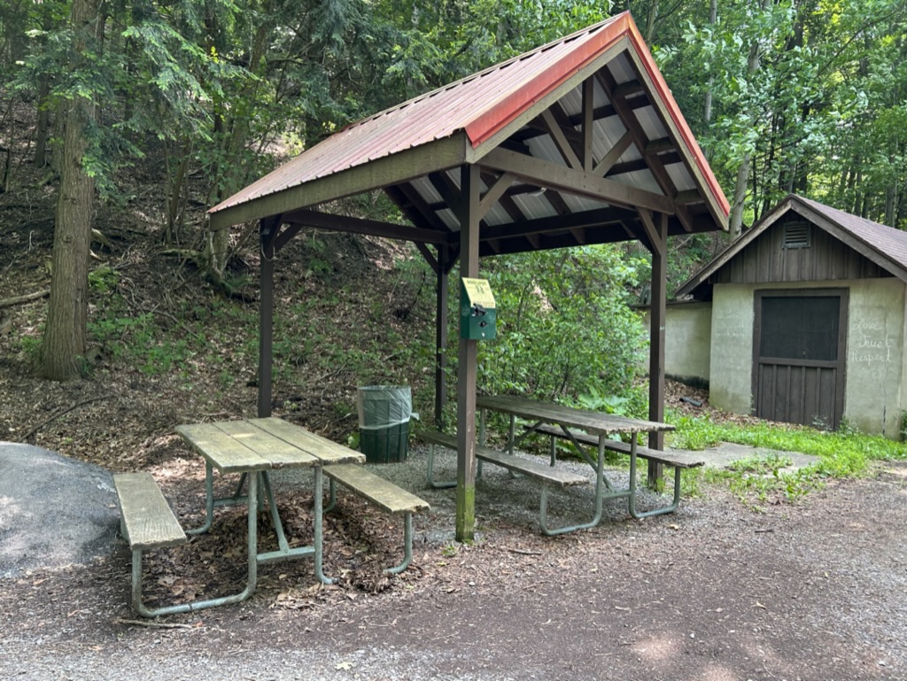A small picnic shelter with two picnic tables at the bottom of a forested hill, sitting next to a concrete builsing with a wooden barn door and gabled roof