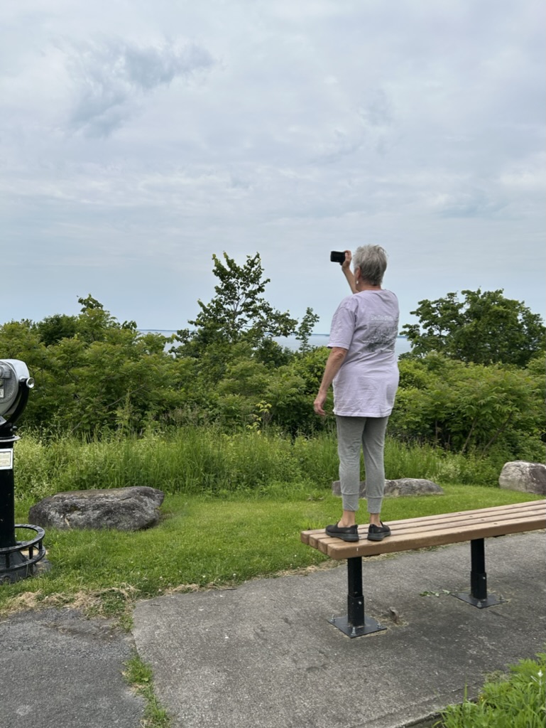 Jill stands on the bench at the overlook in order to see over the bushes to the lake beyond