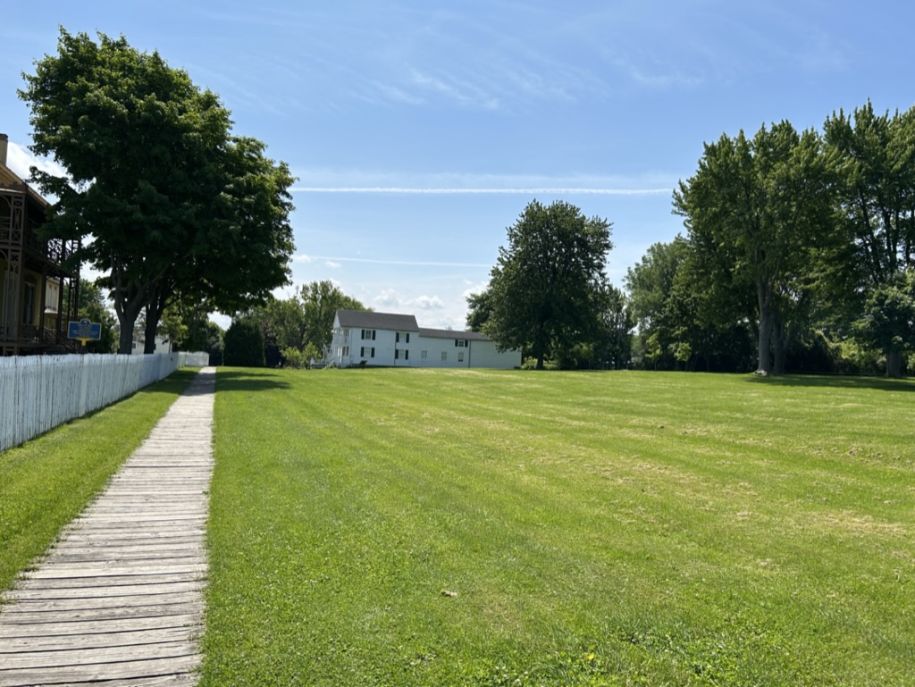 Green field with trees and a white building in the background, with a stone walkway leading along a fence