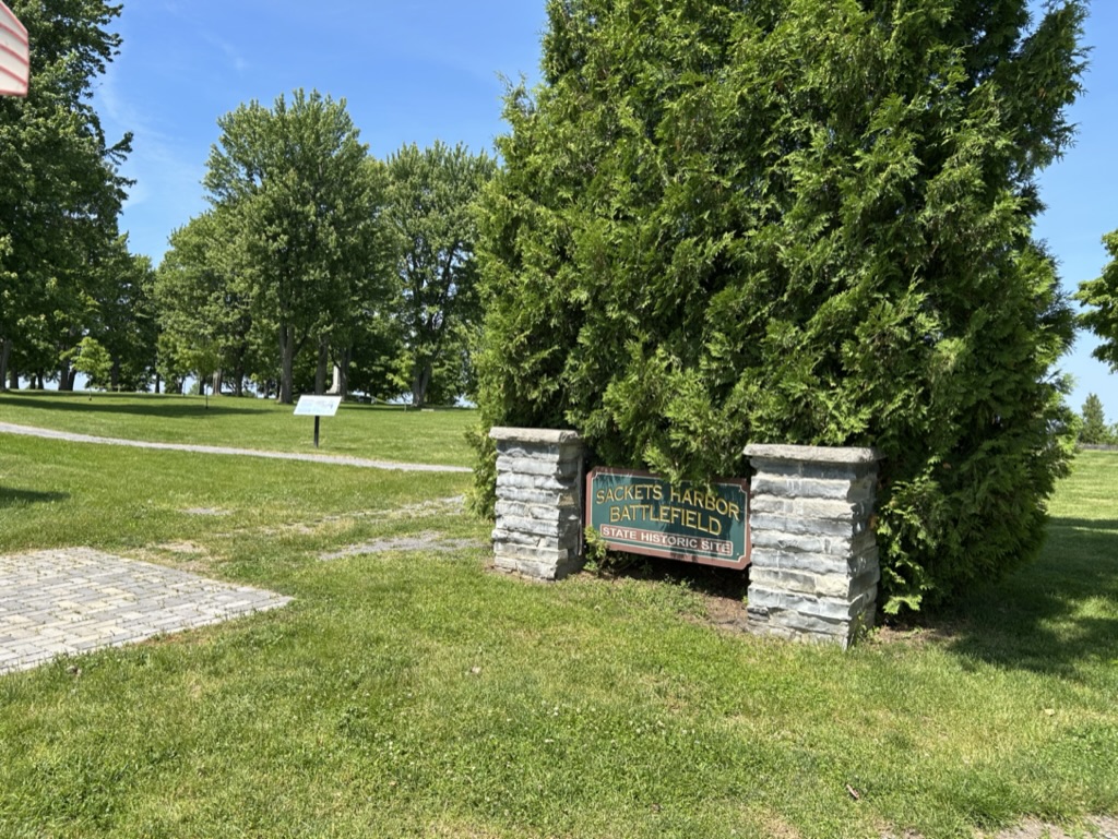 A grassy field with planted trees evenly spaced, and a sign betwen stone supports reading Sackets Harbor Battlefield