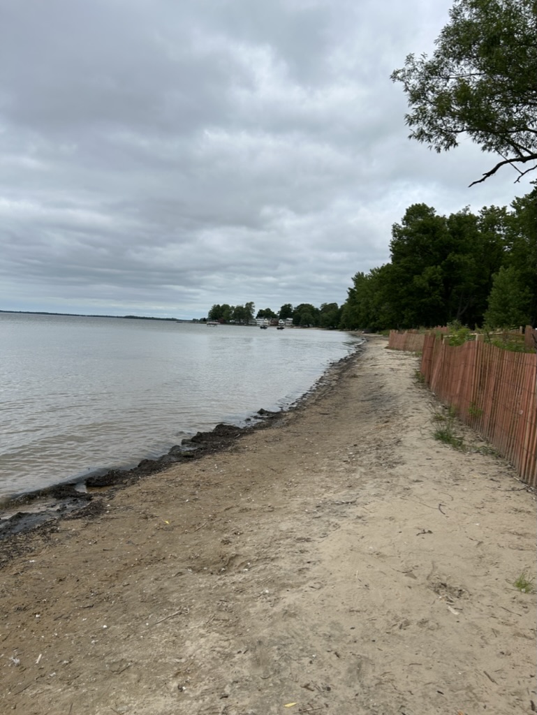 A narrow strip of beach along a lakeshore, enclosed by a reddish wood picket fence. There are trees in the distance and the sky is cloudy.