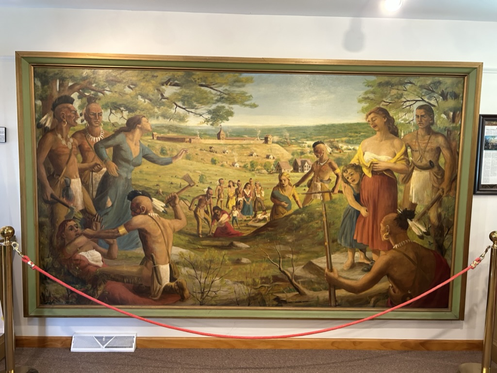 A large painting depicting the village of Fort Plain being attacked by Mohawk tribesmen, who have captured several white women. One woman is trying to escape or plead for her life, one is on the ground being threatened by a Mohawk with a tomahawk, another is standing clutching a child at her side.