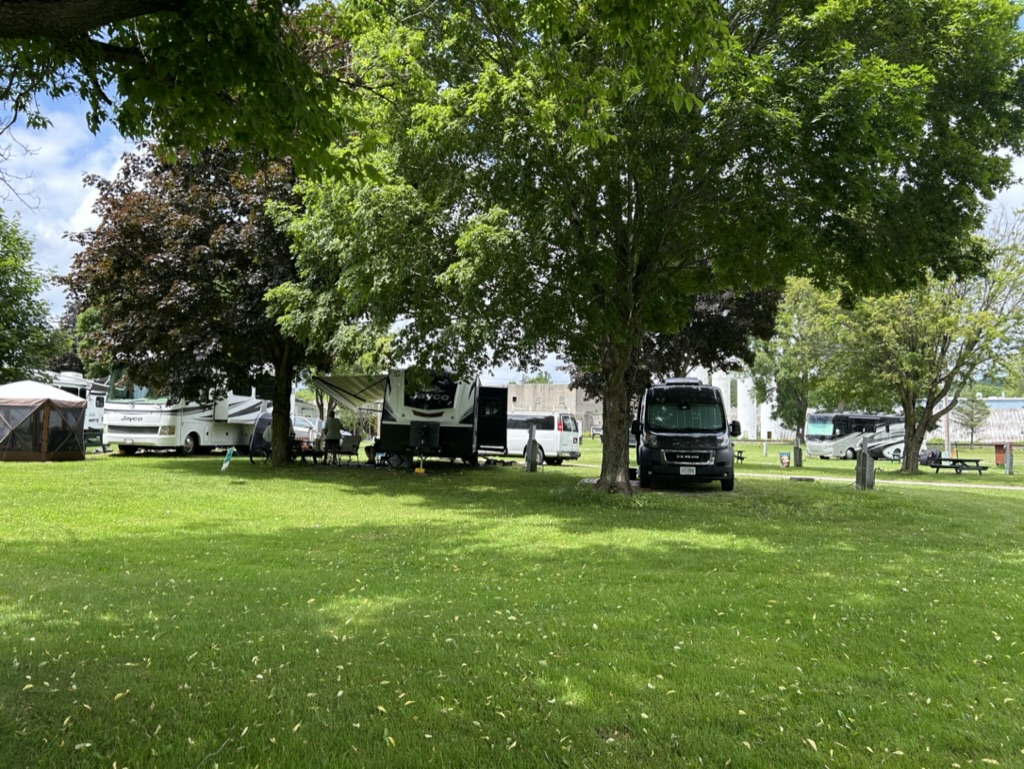 A van and several trailers parked on the grass among tall shade trees