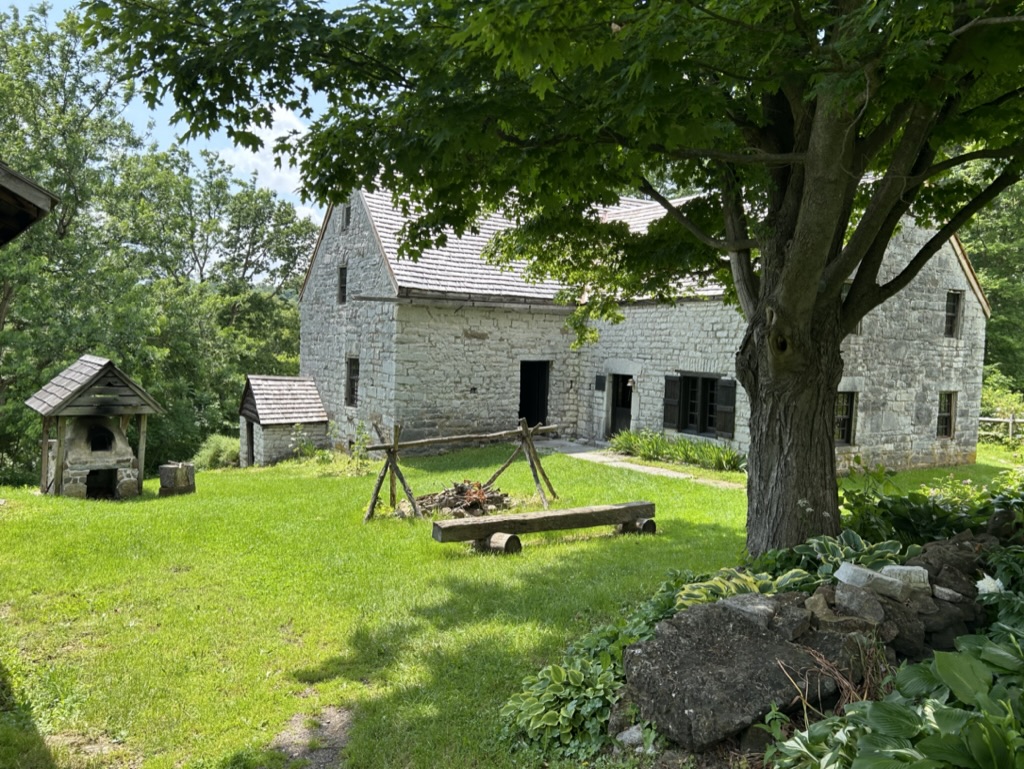 Fort Klock, a large L-shaped white stone and brick building. There a fire pit in front with wooden framework over it to hang cooking pots from, and a low bench made from a split log. To the side is a brick bee-hive shaped oven with a wooden roof built over it