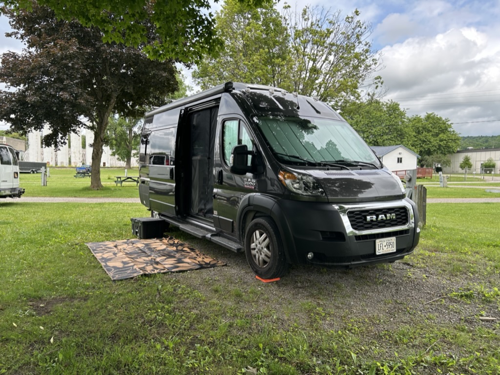 Gray van parked in a grassy site with its sliding door open and an outdoor rug on the ground in front of the door