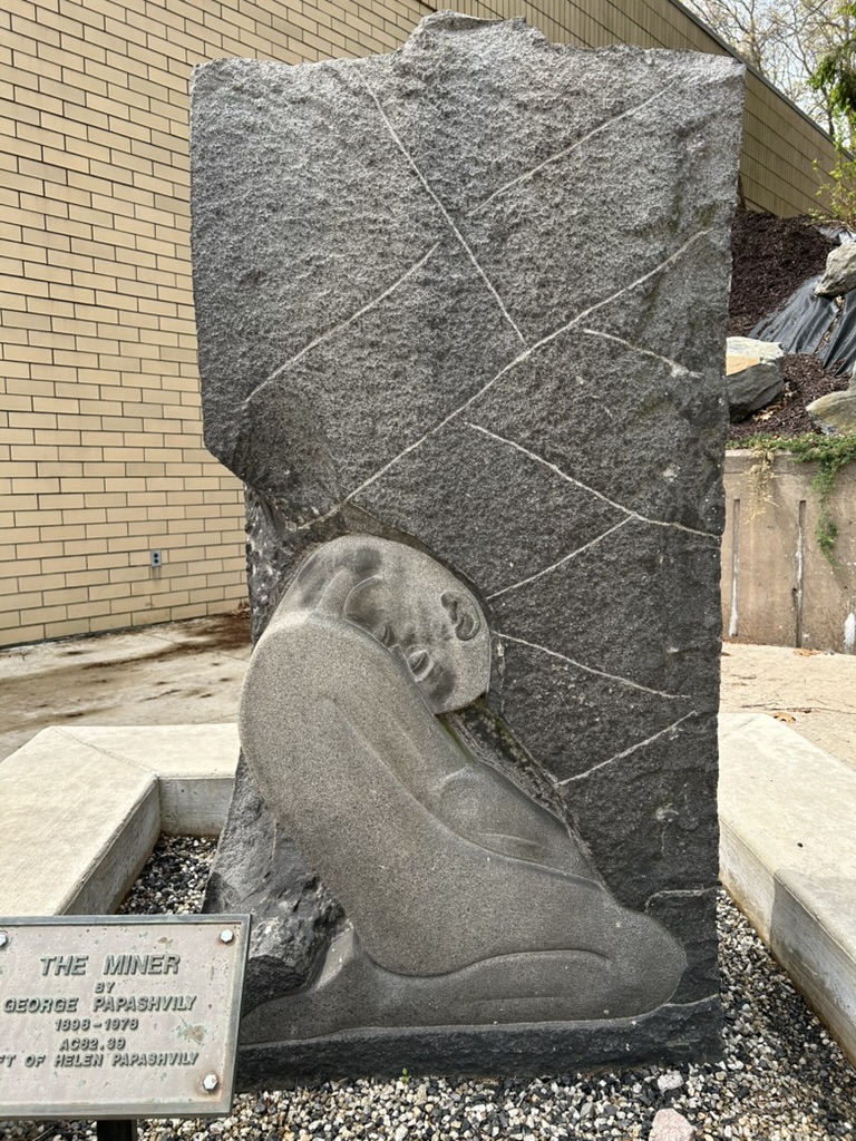 A stone sculpture called The Miner, showing the stylized figure of a man on his knees with his head bent forward onto his chest, being crushed by the weight of stone above him