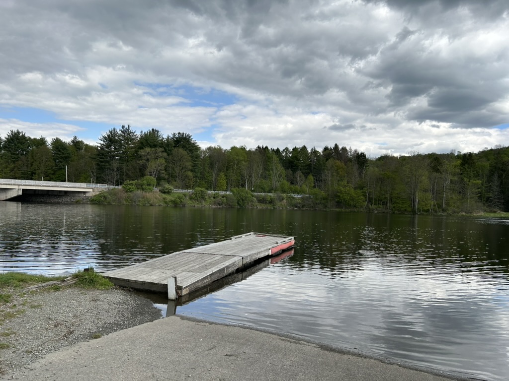 A small wooden dock next to a concrete boat launch on a lake. There is a bridge across the lake to the left, and trees along the opposite shore.