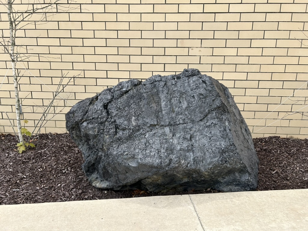 A large boulder of anthracite coal sits on mulch in front of a tan brick building