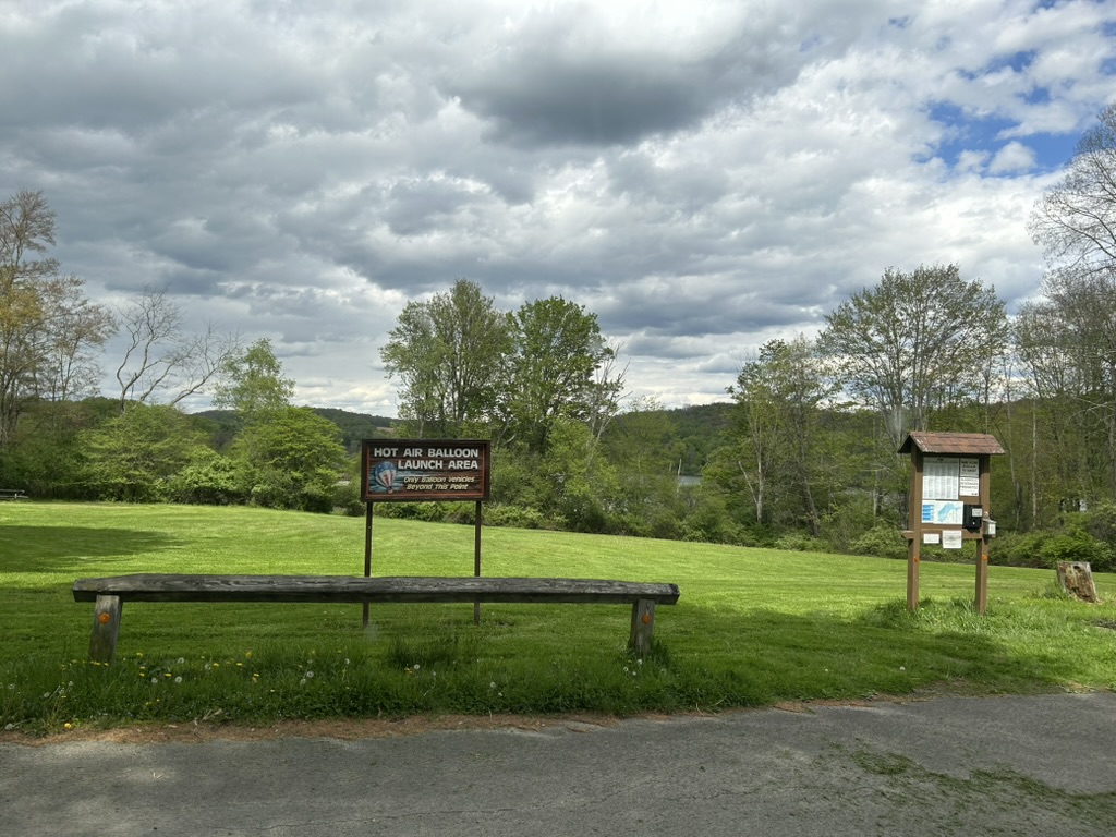 A grassy field under a cloudy sky. In the distance there is a glimpse of a lake beyond the trees. A sign in the foreground reads: Hot Air Balloon Launch Area