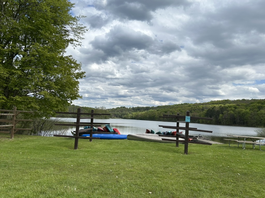 A grassy shoreline with three wooden stands for holding kayaks in front of the lake. There are a number of colorful kayaks pulled up on the short.
