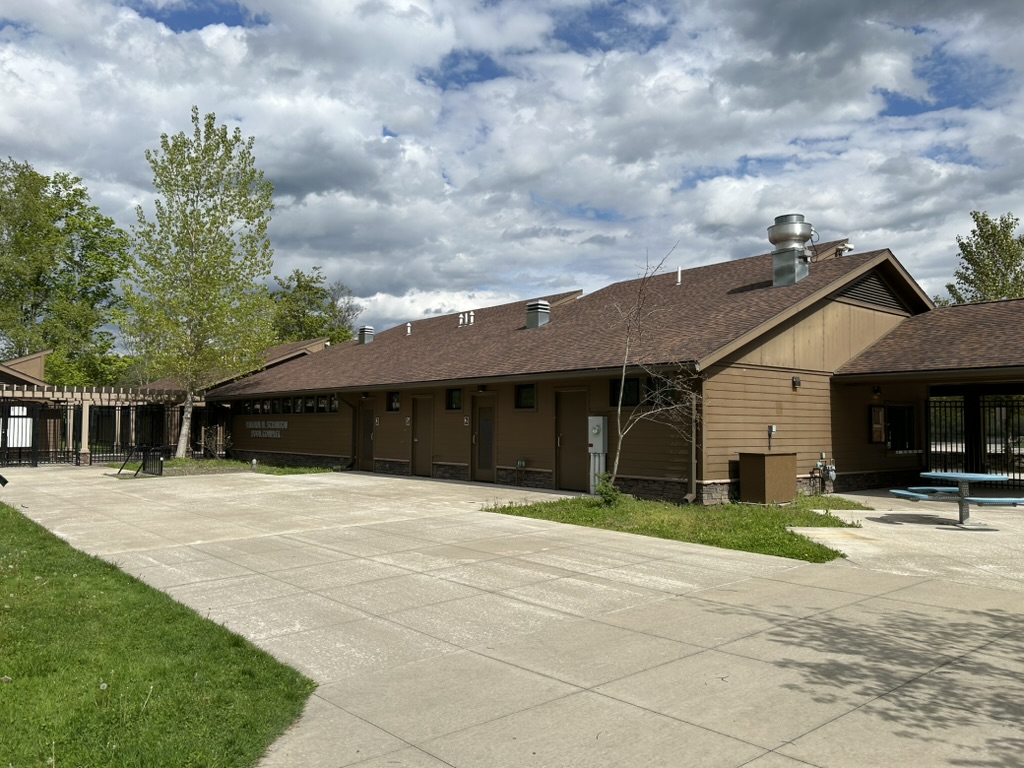 The William W Scranton Pool Complex is a long low brown building painted brown with a row of changing rooms along the side. There is a concrete area in front with a picnic table and chairs at the right side.