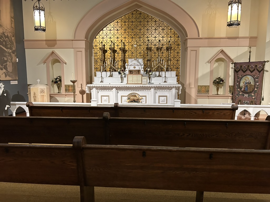 Exhibit of a church. A white painted altar with a tall candles on top sits in an alcove with a gold patterned background. In front are wooden pews