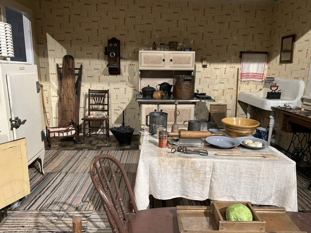 Exhibit of a typical farmhouse kitchen. On the left is a large enameled ice chest. Along the back is an enameled wood stove, some chairs, and a large farmhouse sink. In the front is a large kitchen table with various cooking implements and dishes.