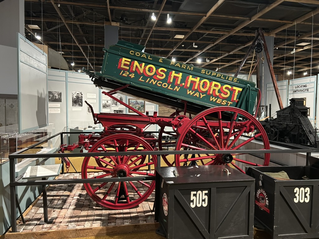 Exhibit of a green coal delivery cart with red wheels. The cart is tipped up so the coal can slide out. On the side it reads, Coal and Farm Supplies, Enos H. Horst, 124 Lincoln Way West