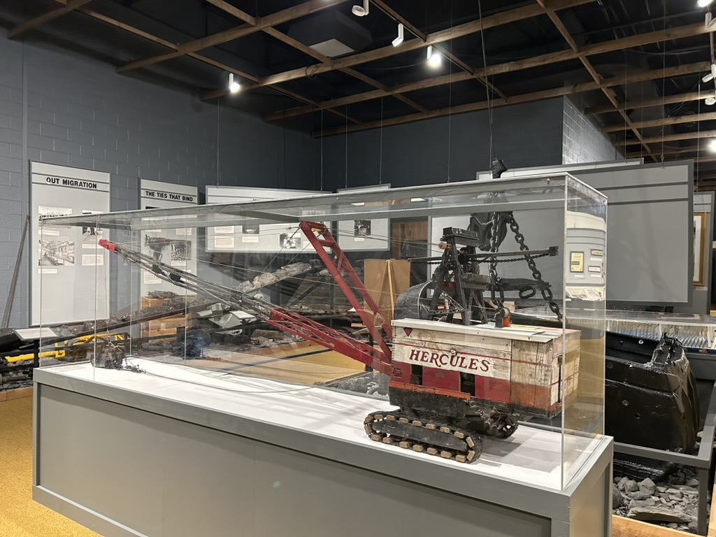 Model of a crane in a large display case. The body of the crane is painted white and red and reads Hercules on the side. It has a long crane arm and continuous track treads.